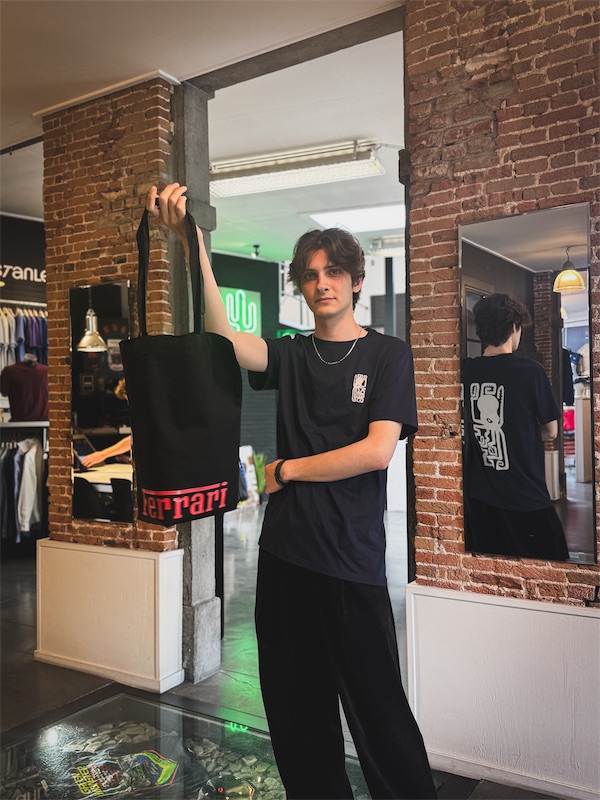 Andrei staat tussen twee bakstenen pilaren in de winkel van PAS PRINT en houdt een zwarte draagtas met rode Ferrari-opdruk omhoog. Zijn donkerblauwe T-shirt heeft een klein wit symbool op de borst en een grotere versie op de rug, zichtbaar in de spiegel.