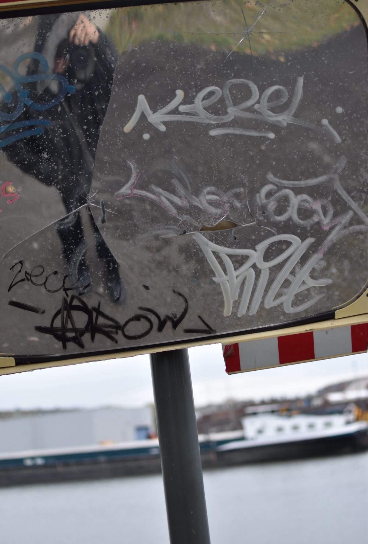 Graffiti and reflections on a weathered traffic mirror, with a waterway and a boat in the background.